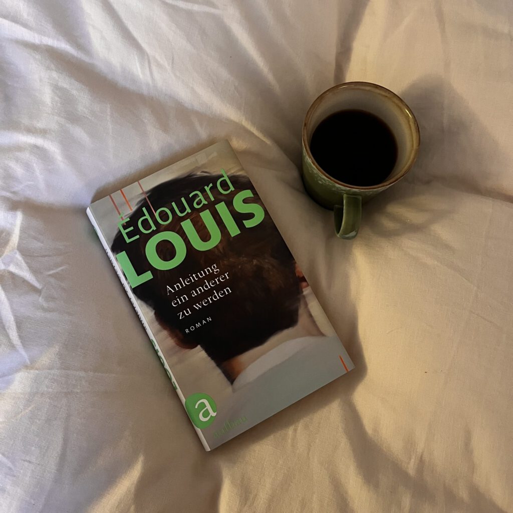 Das Buch Anleitung ein anderer zu werden von Edouard Louis liegt neben einer grünen Tasse mit Kaffee in warmem Licht auf einer beigen Bettdecke.