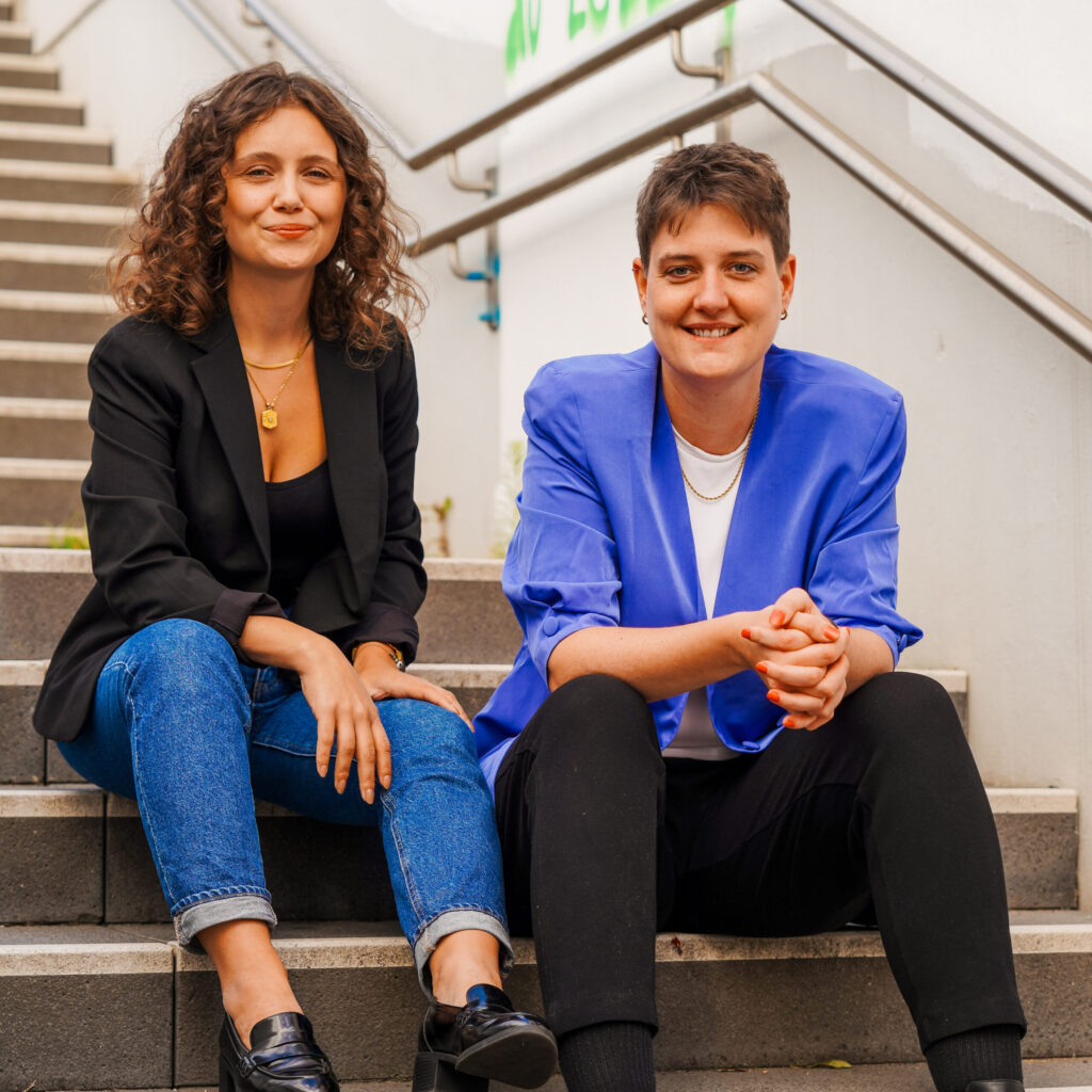 Sina Schmeiter und Lisa Wessel sitzen nebeneinander auf einer Treppe und lächeln in die Kamera.